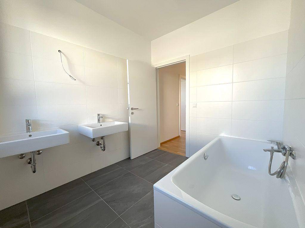 Helles Badezimmer mit Badewanne, zwei Waschbecken und Zugang zu einem weiteren Raum.