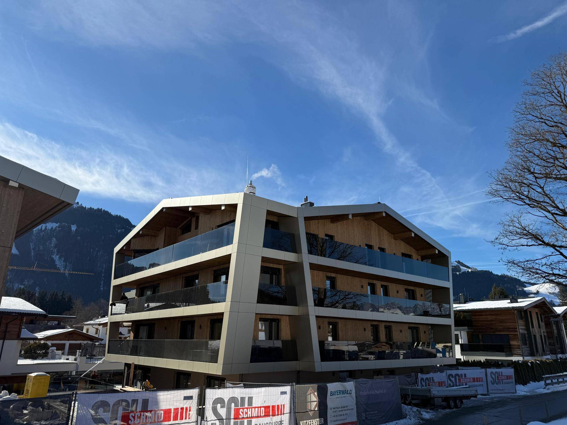 Modernes Apartmentgebäude mit Holz- und Glasfassade im Winter, umgeben von Schnee und Bergen.