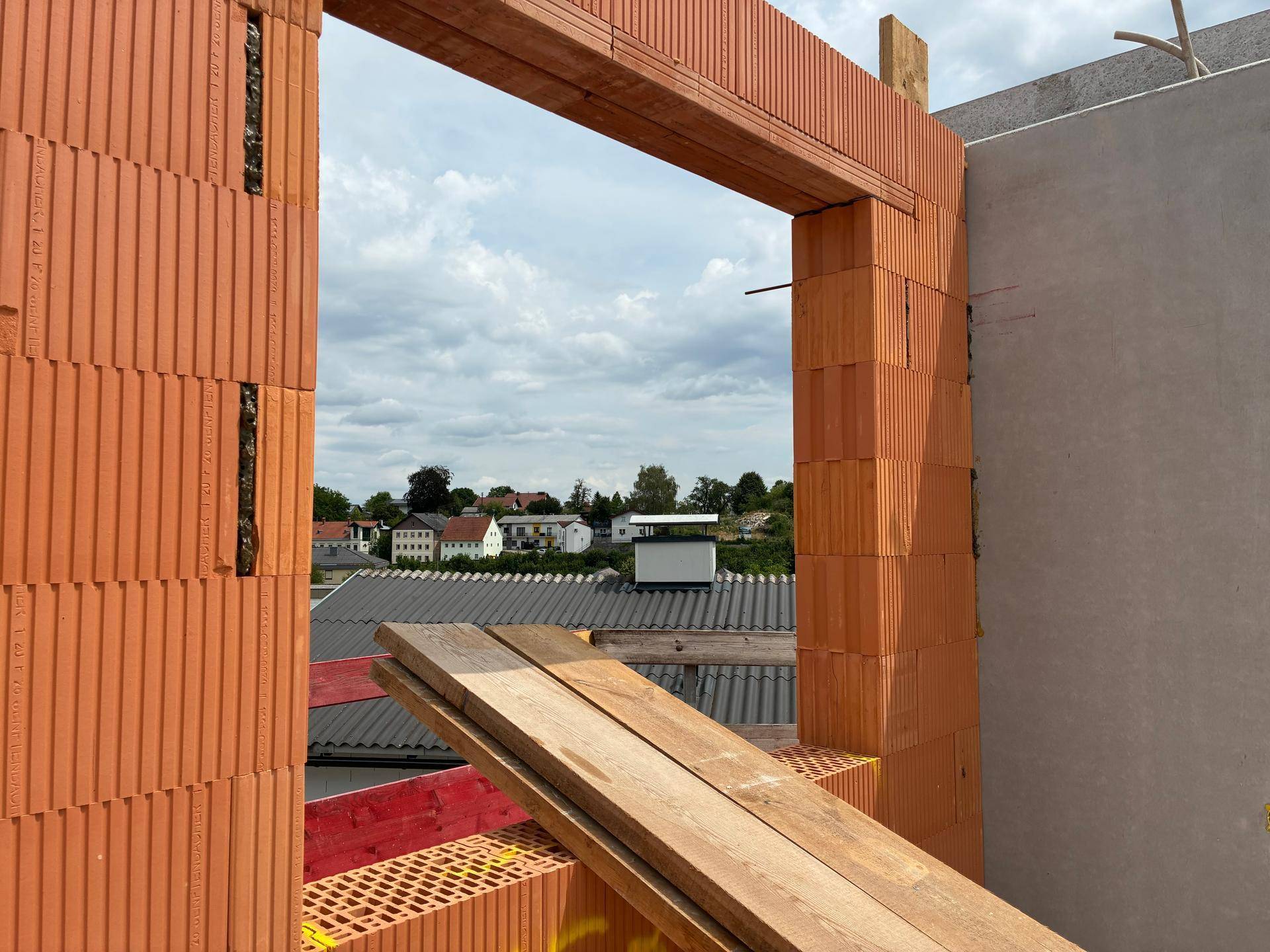 Fensteröffnung in einer Ziegelwand mit Blick auf die umliegende Bebauung und den Himmel.