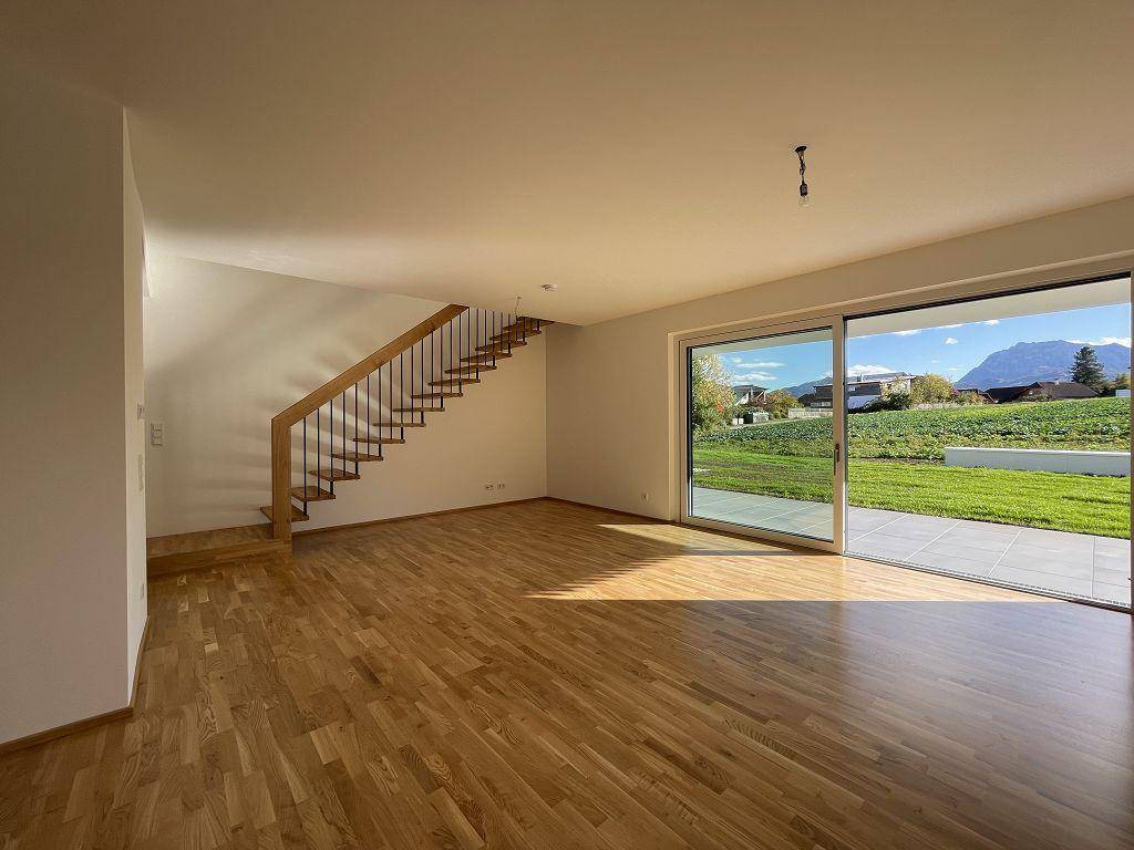 Heller, leerer Wohnbereich mit Holzboden, Treppe und großen Fenstern mit Bergblick und Gartenzugang.