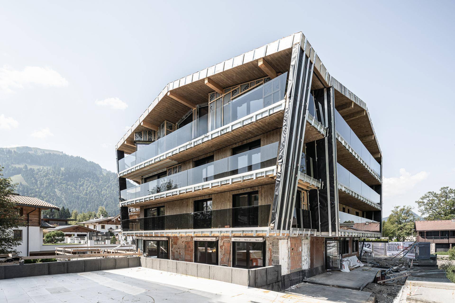 Modernes Gebäude im Bau mit Holzverkleidung, Glasbalkonen und Blick auf die umliegenden Berge.
