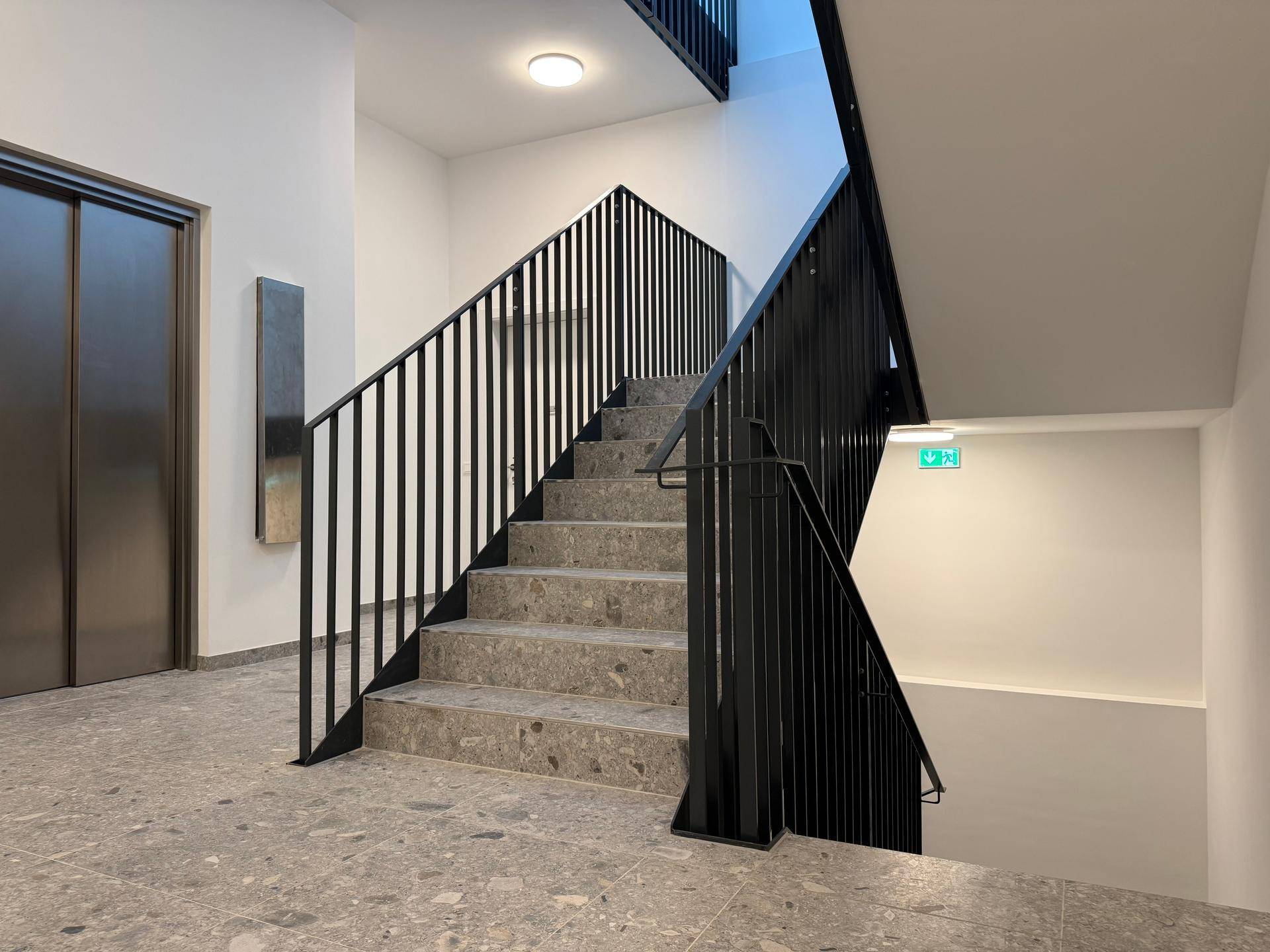 Helles Treppenhaus mit modernem Geländer und Zugang zum Aufzug in einem Neubau.
