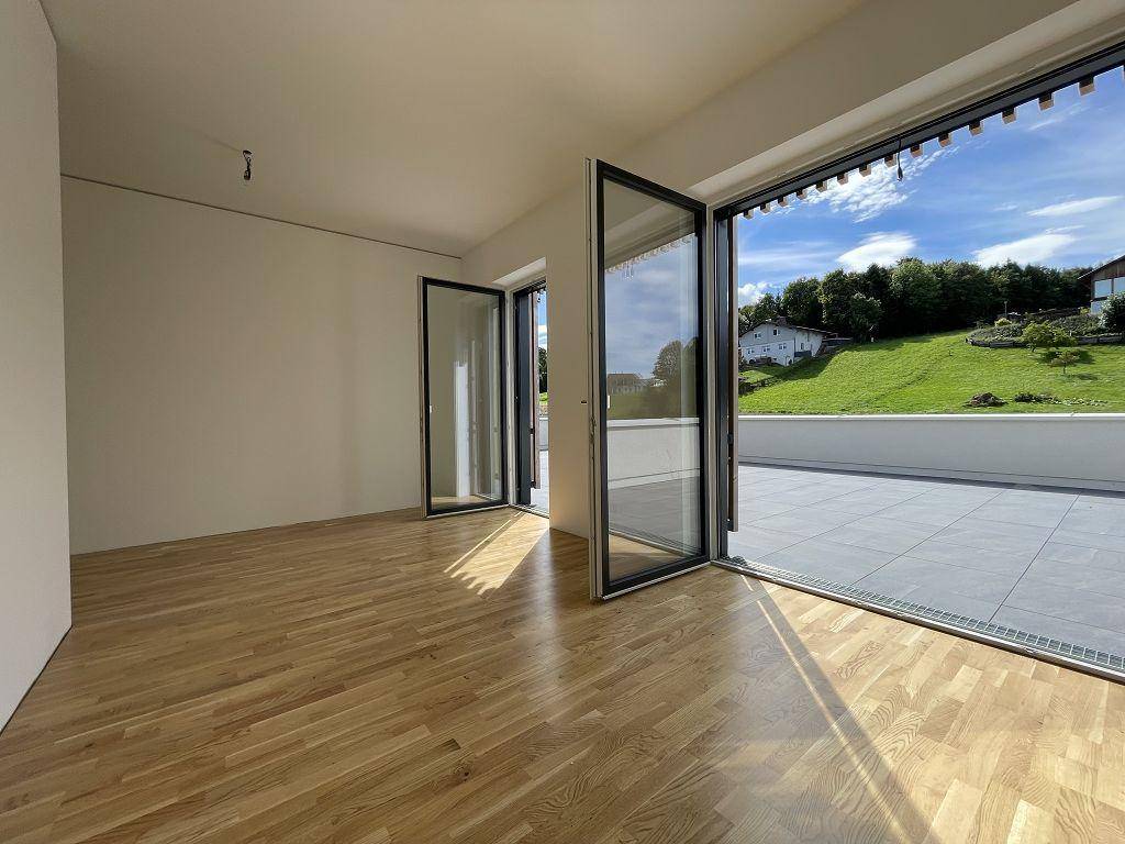 Heller, leerer Raum mit Holzboden und großen Glastüren, die auf eine geflieste Terrasse führen.