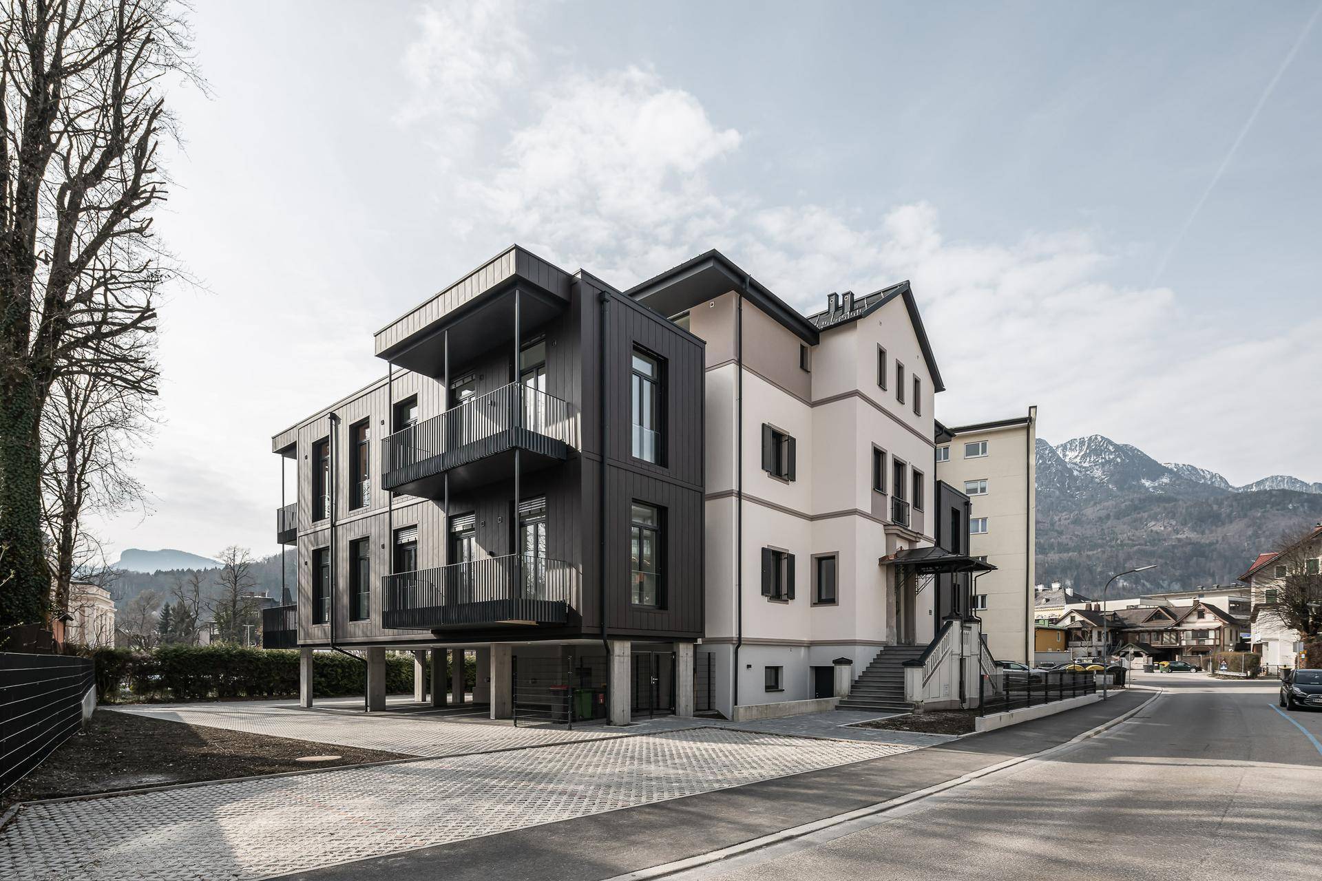 Moderne Architektur eines Neubaus mit dunklen Balkonen und heller Fassade vor Bergkulisse.