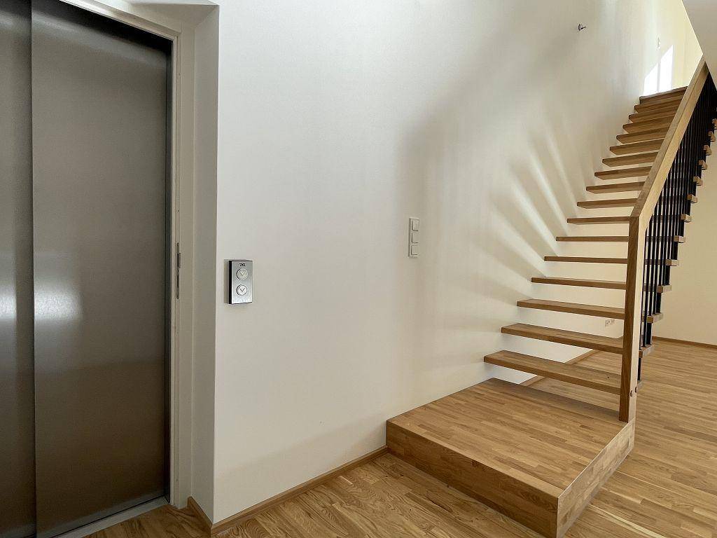 Heller Flur mit Holzboden, moderner Holztreppe und einem Aufzug mit Edelstahltür.