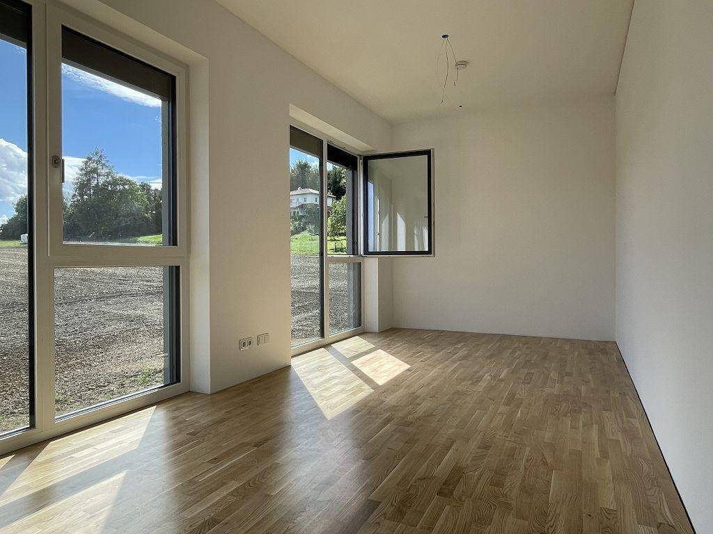 Heller, leerer Raum mit Holzboden und großen Fenstern, die viel Tageslicht hereinlassen.