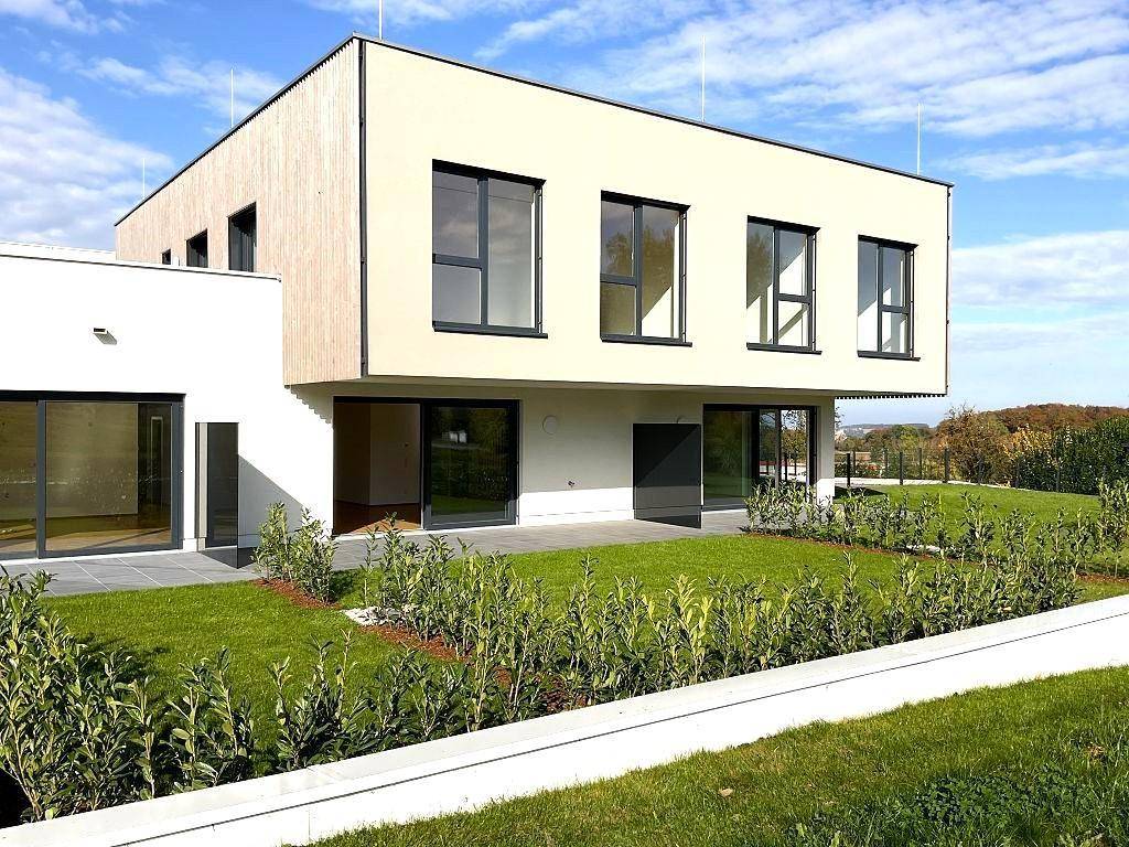 Modernes Neubaugebäude mit weißer Fassade, Holzverkleidung und großen Fenstern, umgeben von Grünflächen.