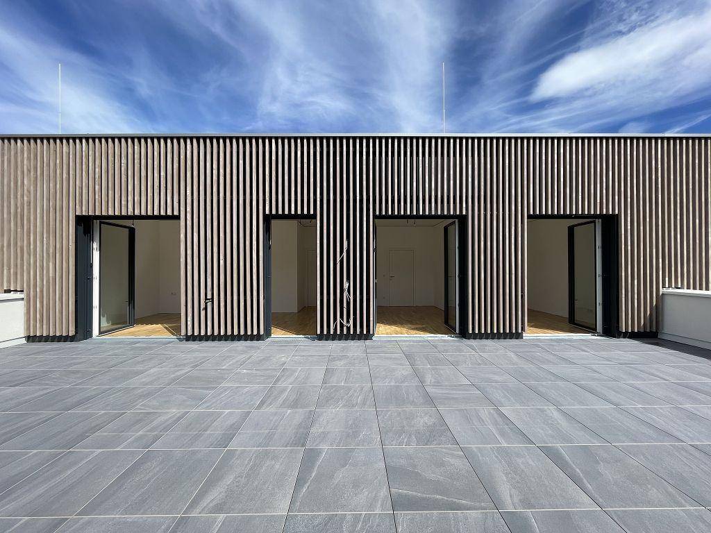 Moderne Holzfassade mit mehreren Glastüren, die auf eine weitläufige, geflieste Terrasse führen.