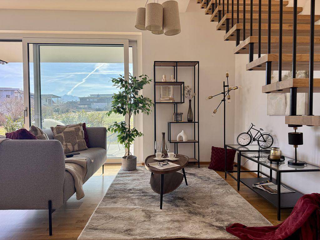 Gemütlicher Wohnbereich mit Sofa, Teppich und Blick durch große Fenster ins Grüne.