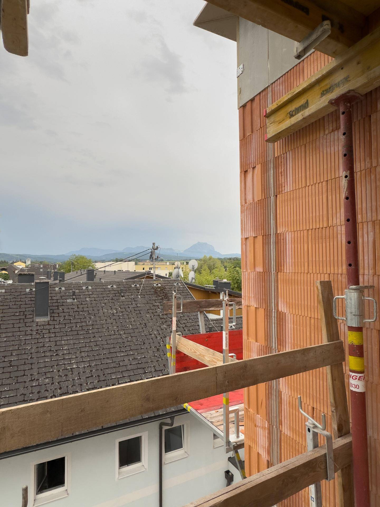 Blick von der Baustelle eines Neubaus über Dächer und die umliegende Berglandschaft unter bewölktem Himmel.