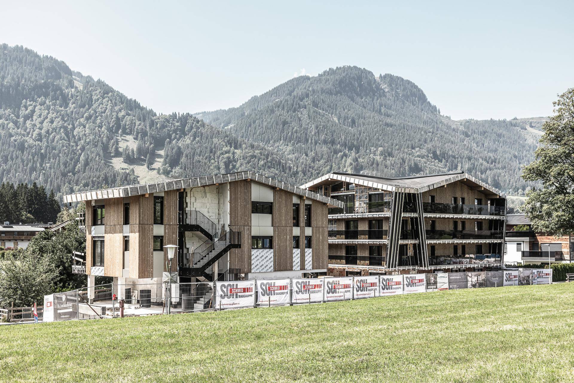Zwei moderne Apartmentgebäude im Bau, eingebettet in eine grüne Landschaft mit Bergen im Hintergrund.