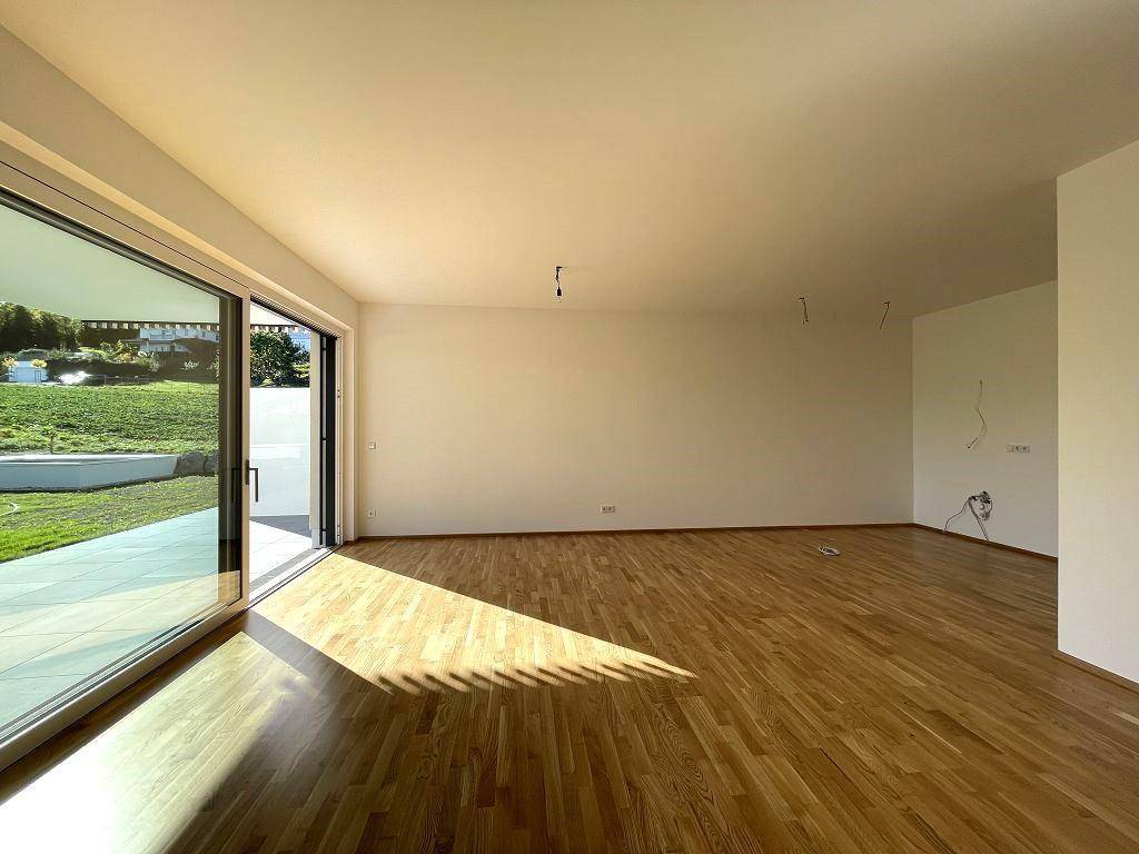 Heller, leerer Wohnbereich mit Holzboden, großen Fenstern und Zugang zu einer Terrasse mit Garten.