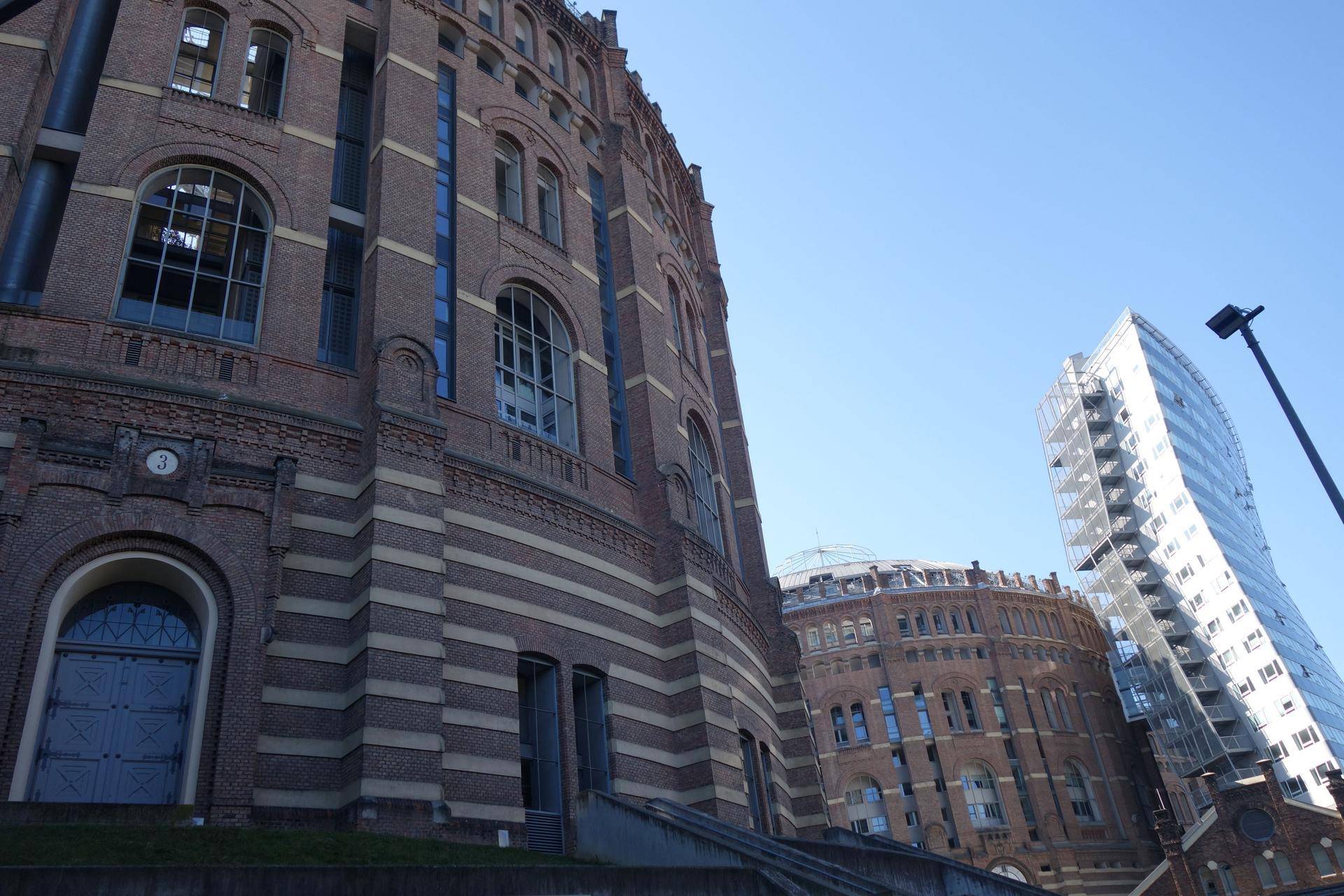 Historische Gasometer-Gebäude aus Ziegelstein neben moderner Glasarchitektur unter blauem Himmel.