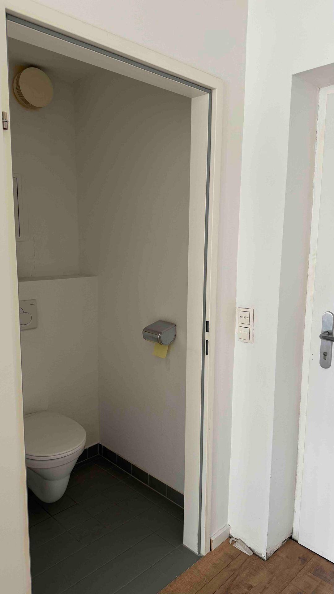 Kleines Gäste-WC mit einfacher Toilette und dunklen Bodenfliesen, ausgestattet mit Abluftsystem.