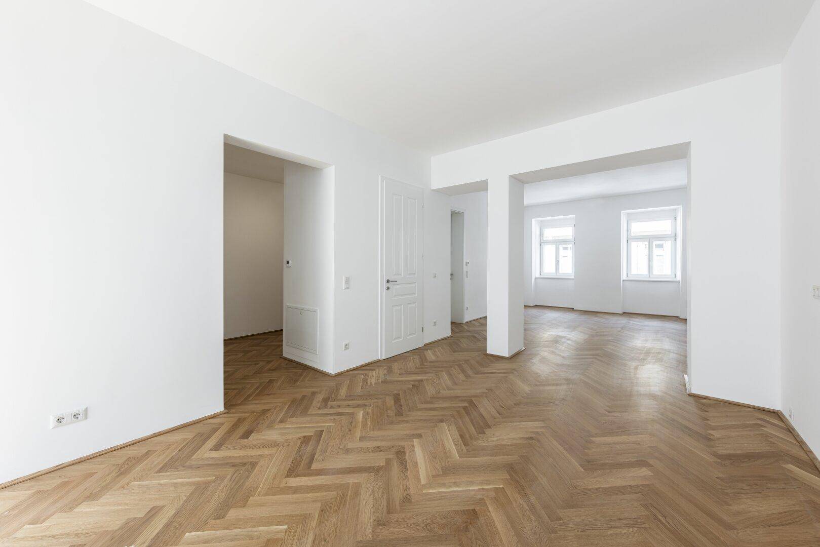 Geräumiger, heller Raum mit Fischgrätparkettboden und mehreren Türöffnungen.