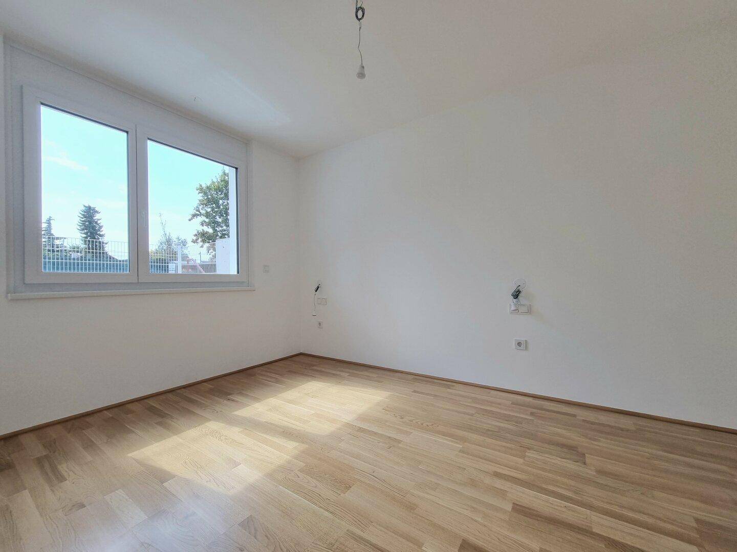 Helles Schlafzimmer mit Parkettboden und großem Fenster, das viel Tageslicht hereinlässt.