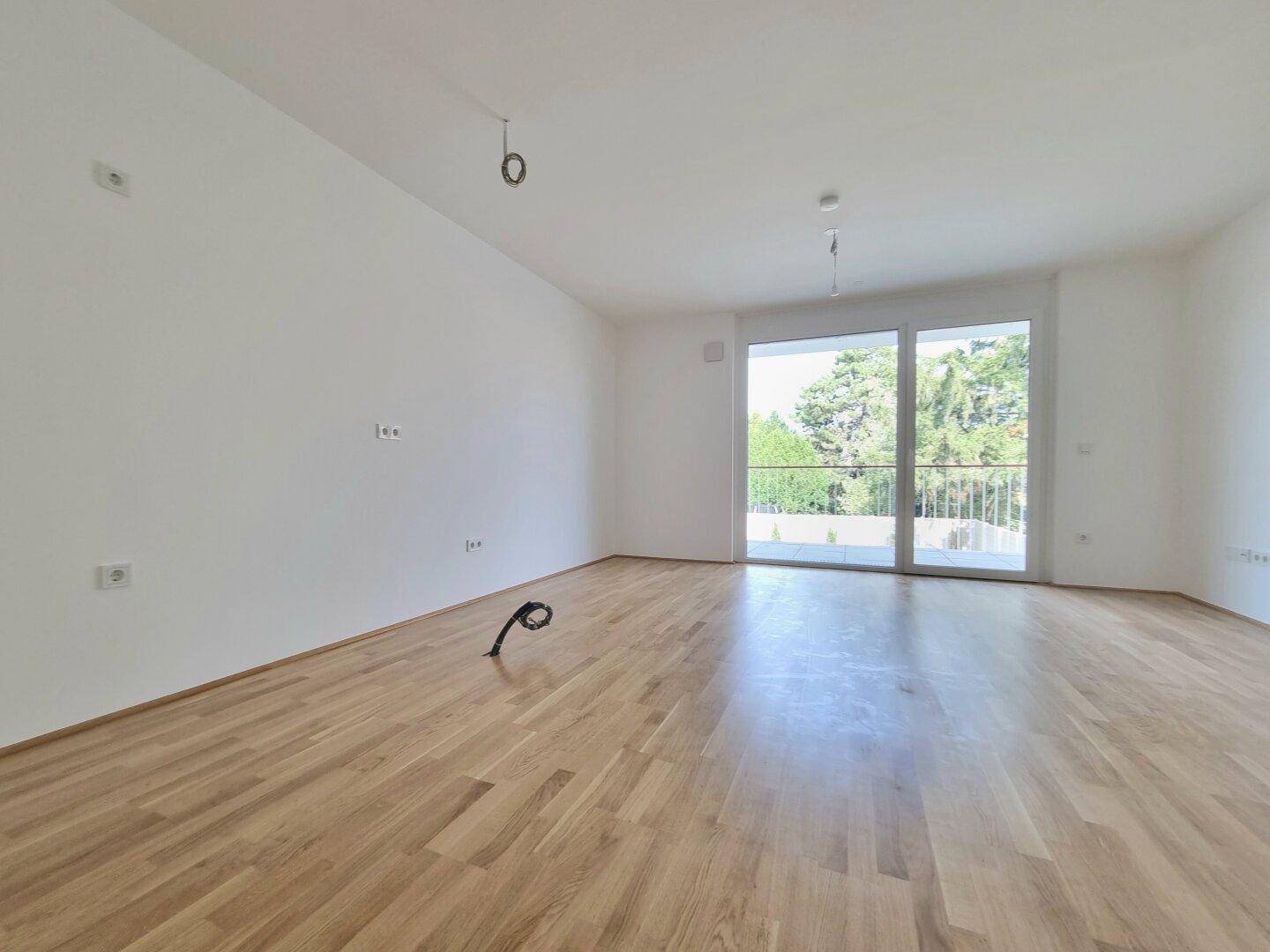 Helles, leeres Wohnzimmer mit Holzboden und großer Glasschiebetür, die zum Balkon führt.
