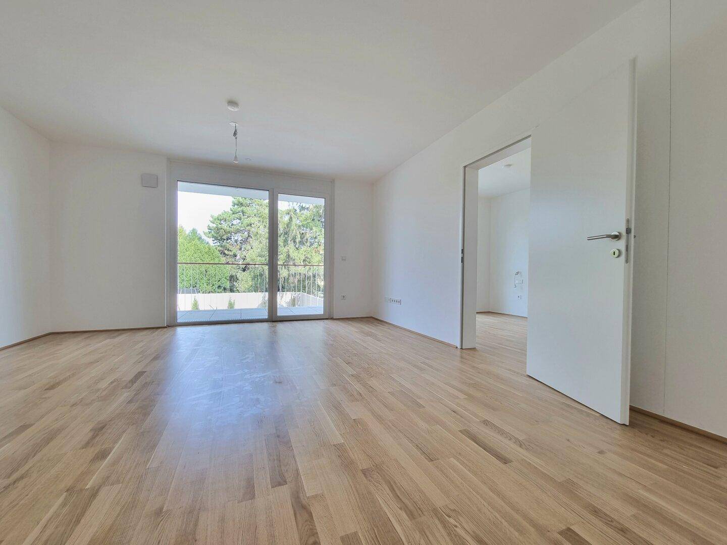 Geräumiges Wohnzimmer mit hellem Holzboden, weißen Wänden und offenem Zugang zu einem weiteren Raum.