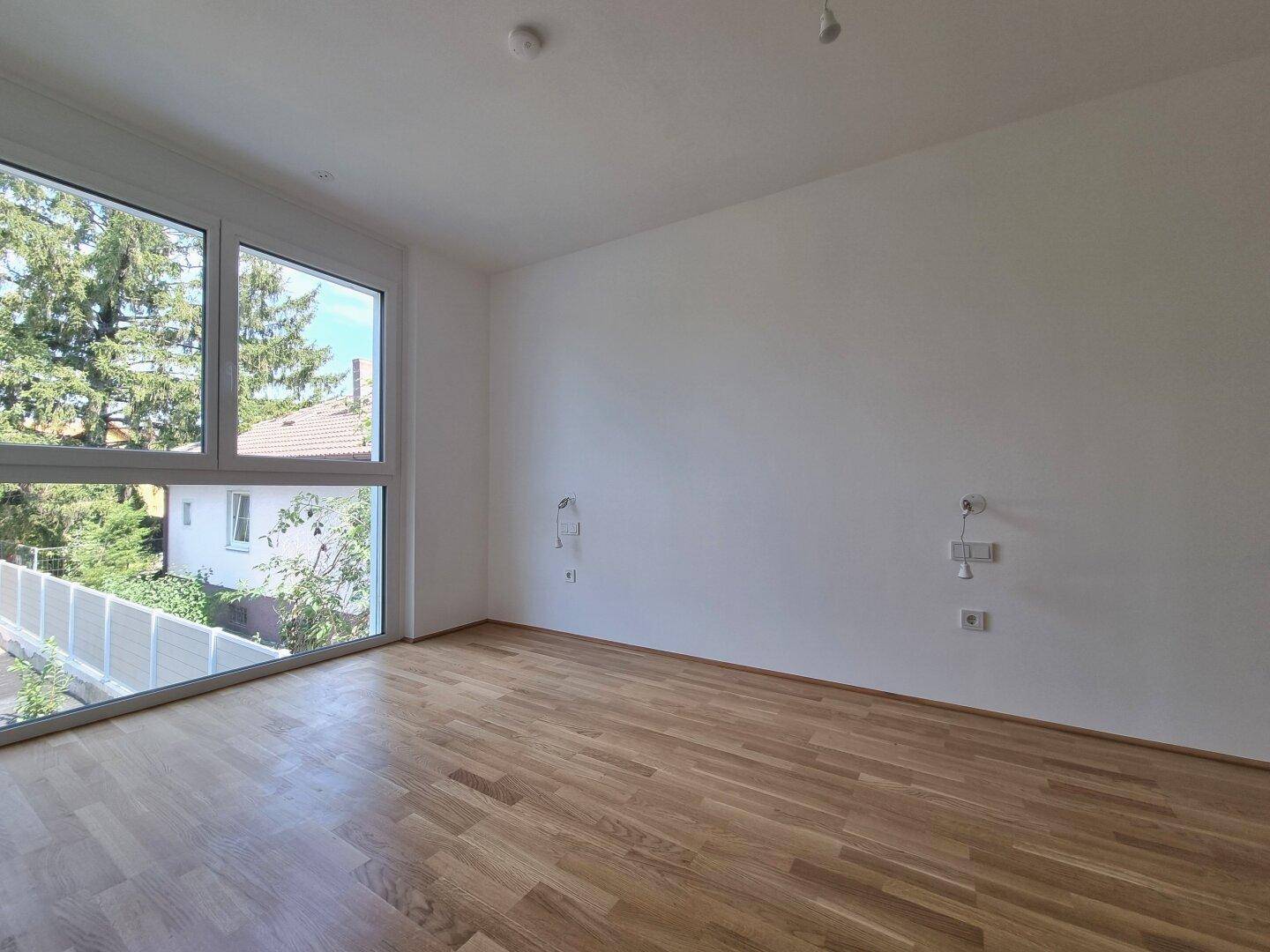 Lichtdurchflutetes Zimmer mit Holzboden und großen Eckfenstern, die viel Tageslicht hereinlassen.