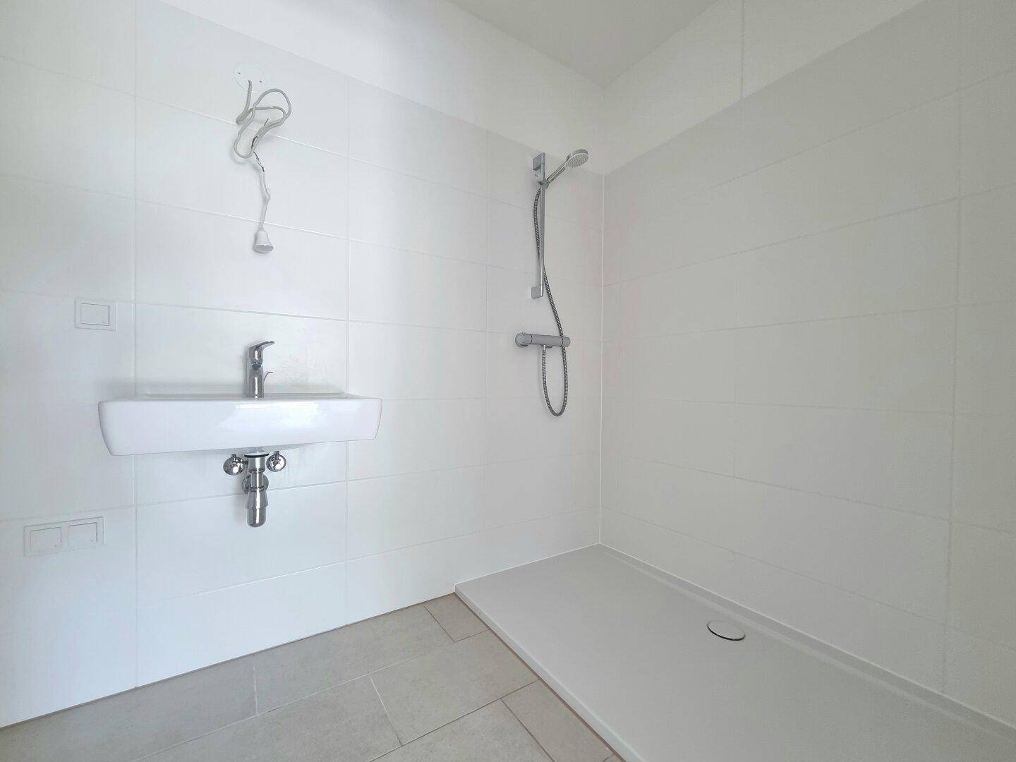Modernes Badezimmer mit weißen Fliesen, wandmontiertem Waschbecken und ebenerdiger Dusche.