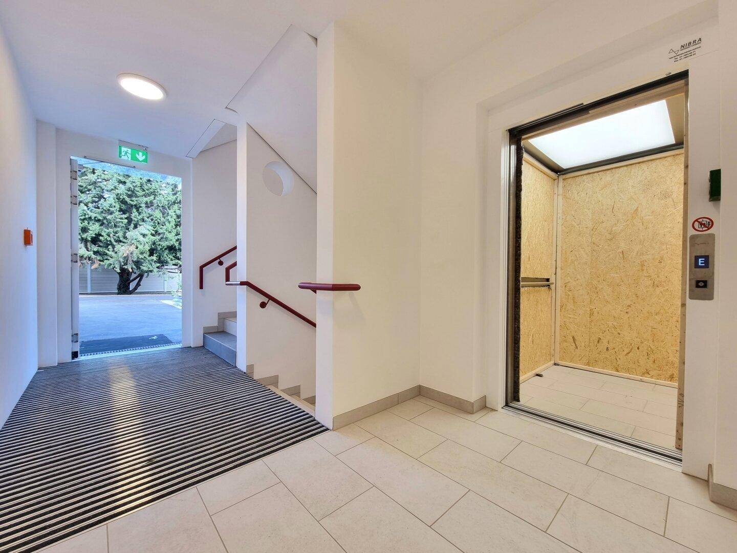 Heller Eingangsbereich mit Rampe, Treppe und modernem Aufzug für barrierefreien Zugang im Gebäude.