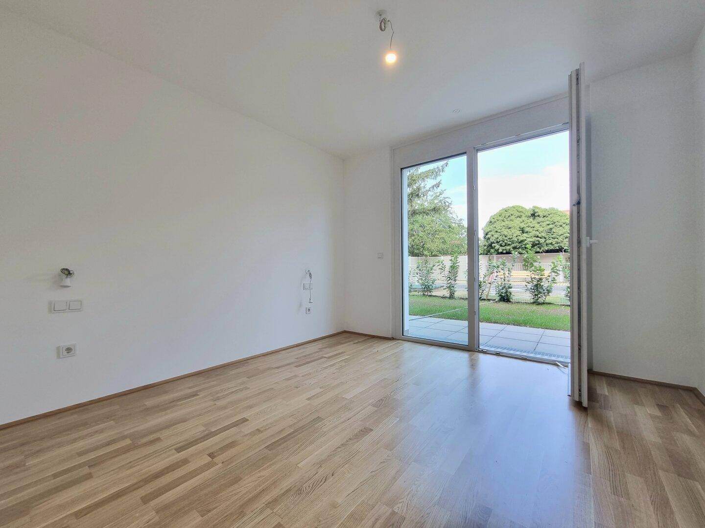 Leeres Wohnzimmer mit Parkettboden und breiter Glasfront zum privaten Garten.