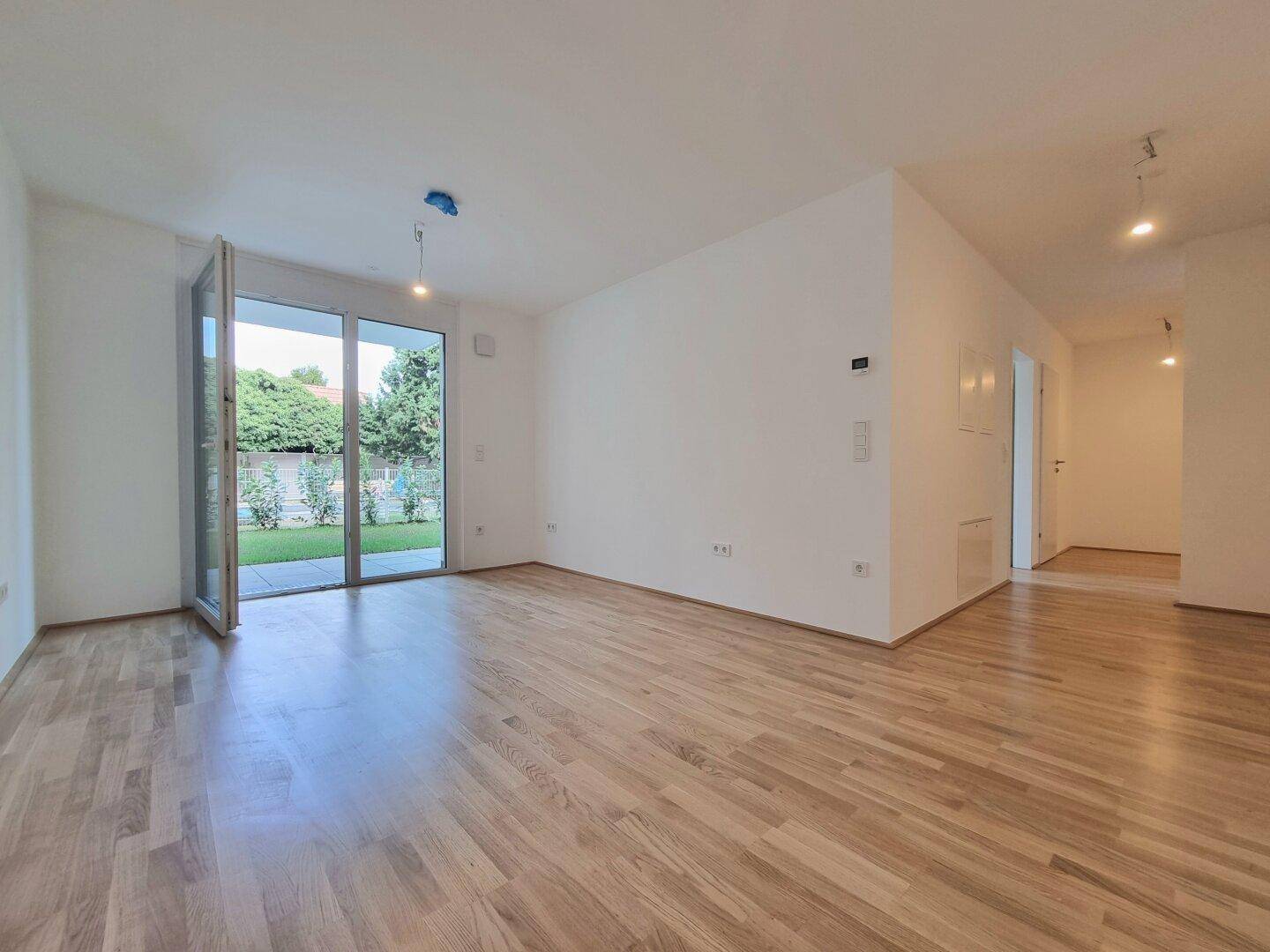 Helles, leeres Wohnzimmer mit Holzboden und Zugang zu einem kleinen Gartenbereich.