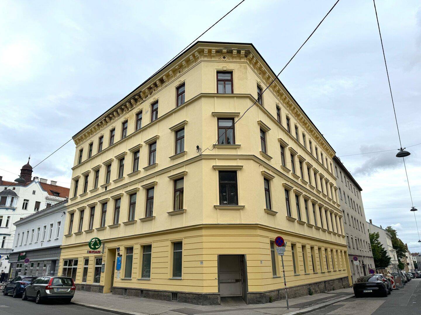 Historisches Eckgebäude mit gelber Putzfassade und vielen Fenstern in urbaner Umgebung.