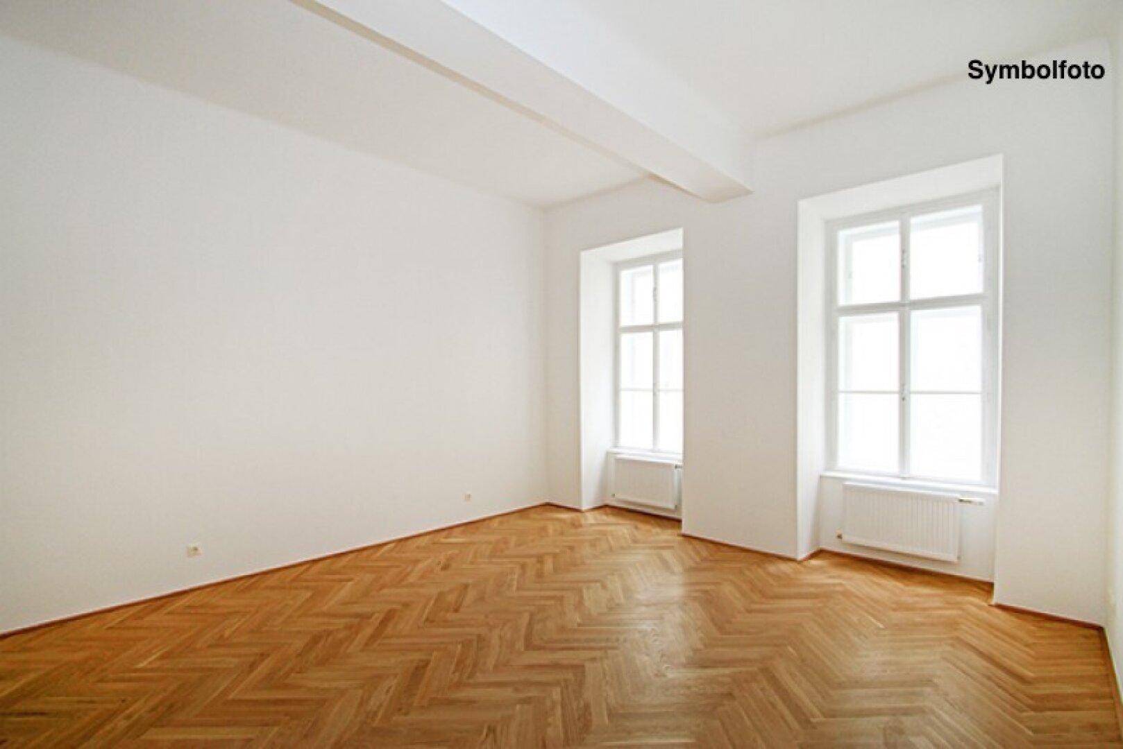 Geräumiger, heller Raum mit Fischgrätparkett, großen Fenstern und Heizkörpern.