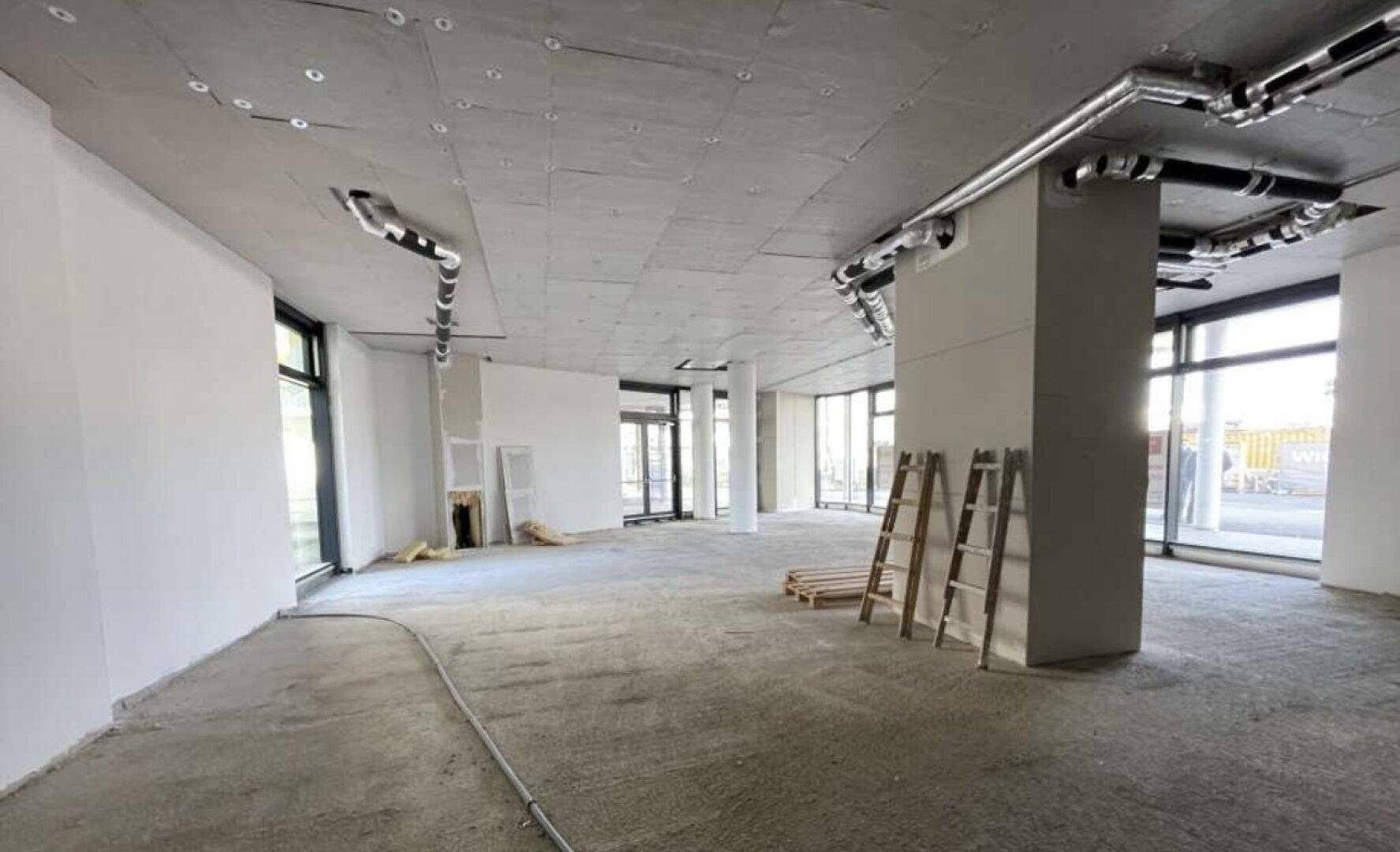 Geräumiger Gewerberaum im Rohbau mit sichtbaren Deckeninstallationen und großen Fensterfronten.