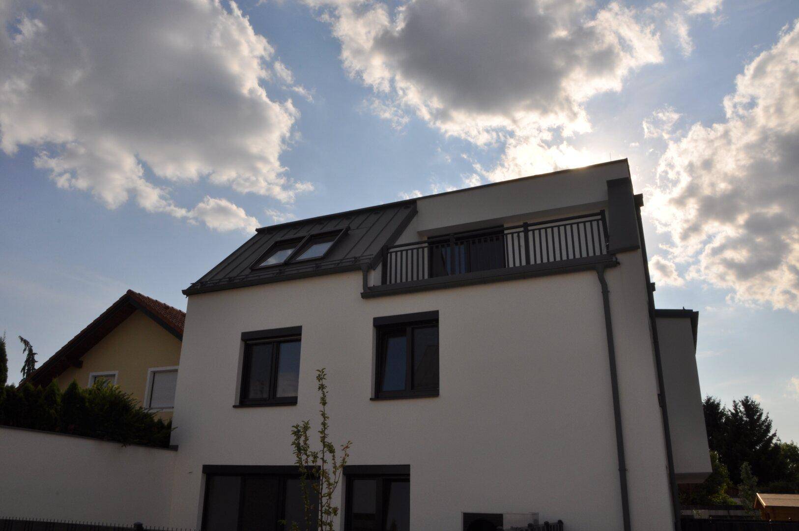 Ansicht des modernen Wohnhauses von der Seite mit Balkon und dunklem Geländer unter sonnigem Himmel.