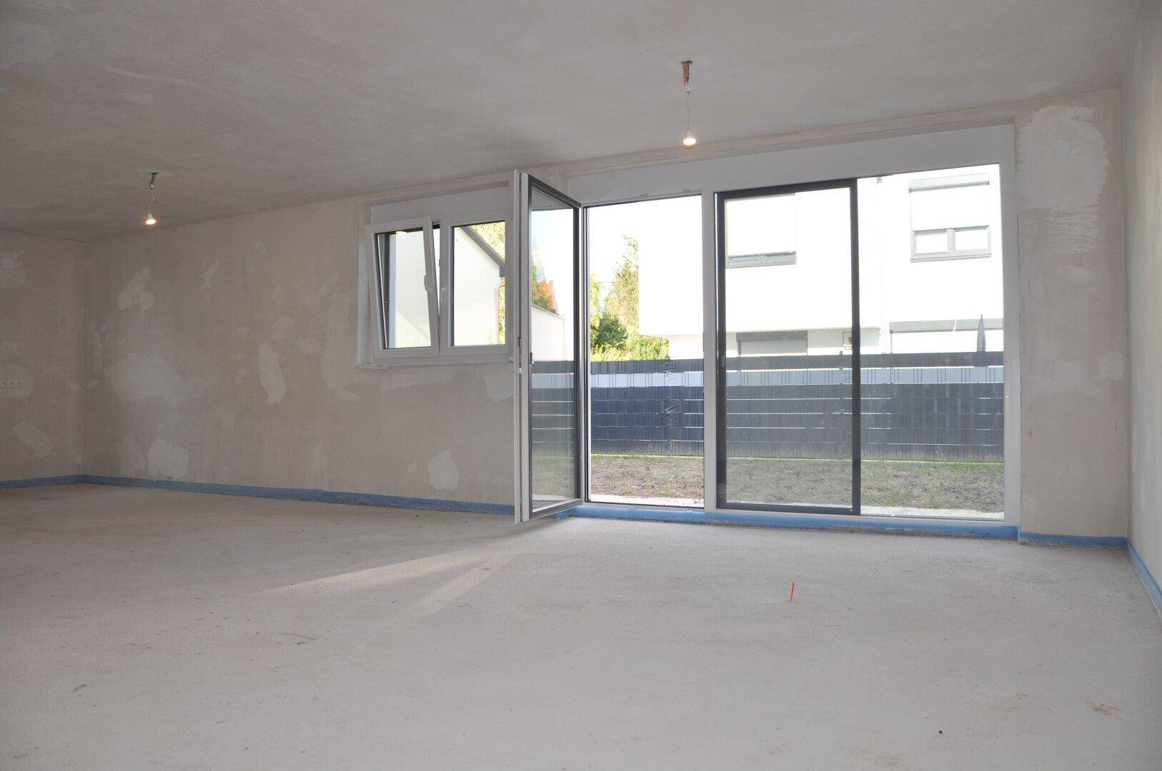 Unfertiges Wohnzimmer mit großen Fenstern und Zugang zum Außenbereich, bereit für den Ausbau.