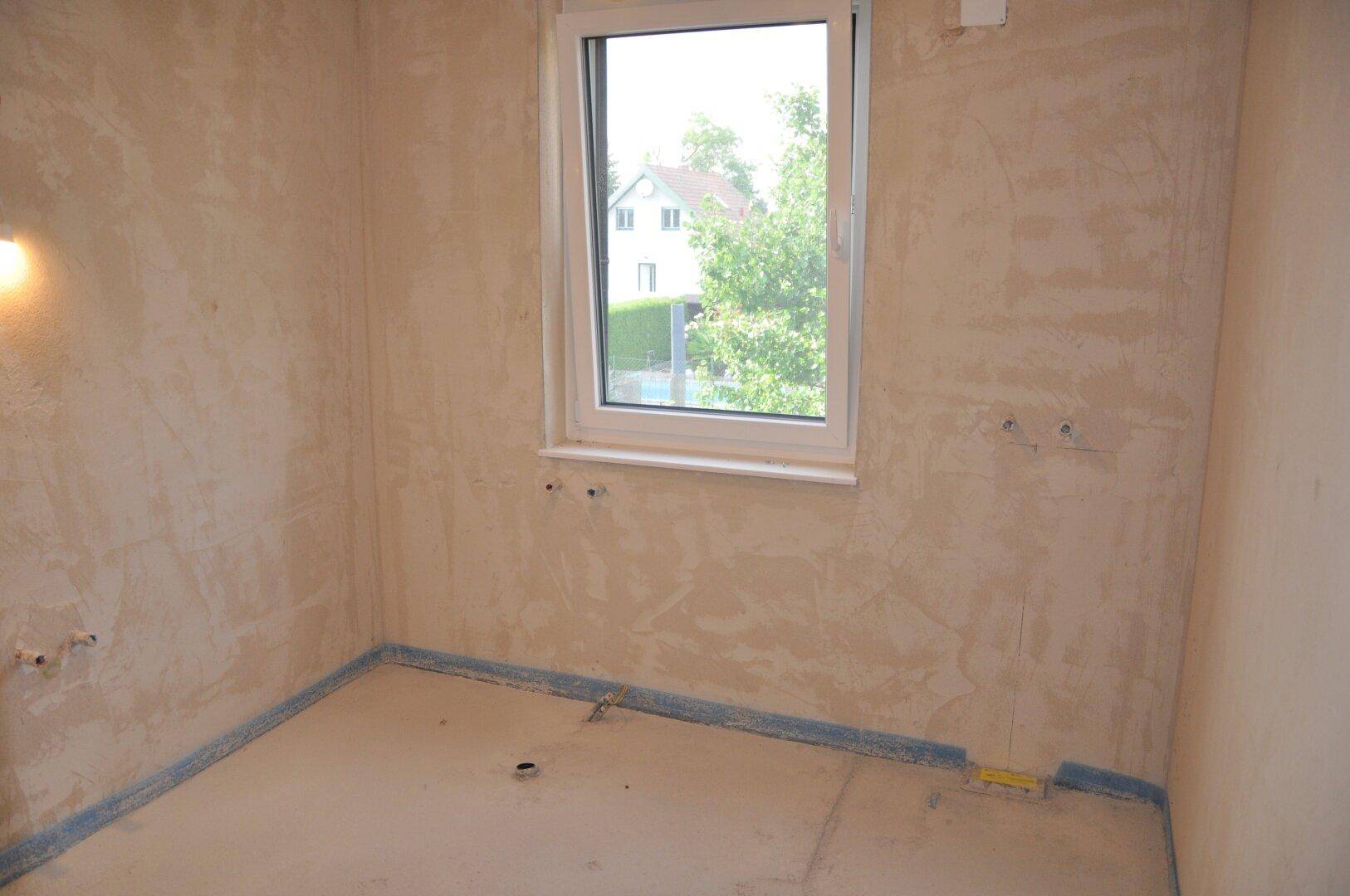 Unfertiger Raum mit Fenster und vorbereiteten Anschlüssen, ideal für ein Badezimmer oder einen Hauswirtschaftsraum.