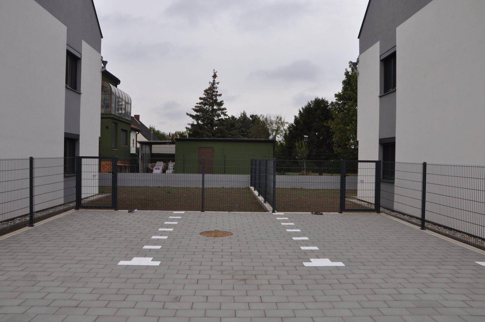Gepflasterter Parkplatz zwischen zwei Reihenhäusern mit Markierungen und Zaun.