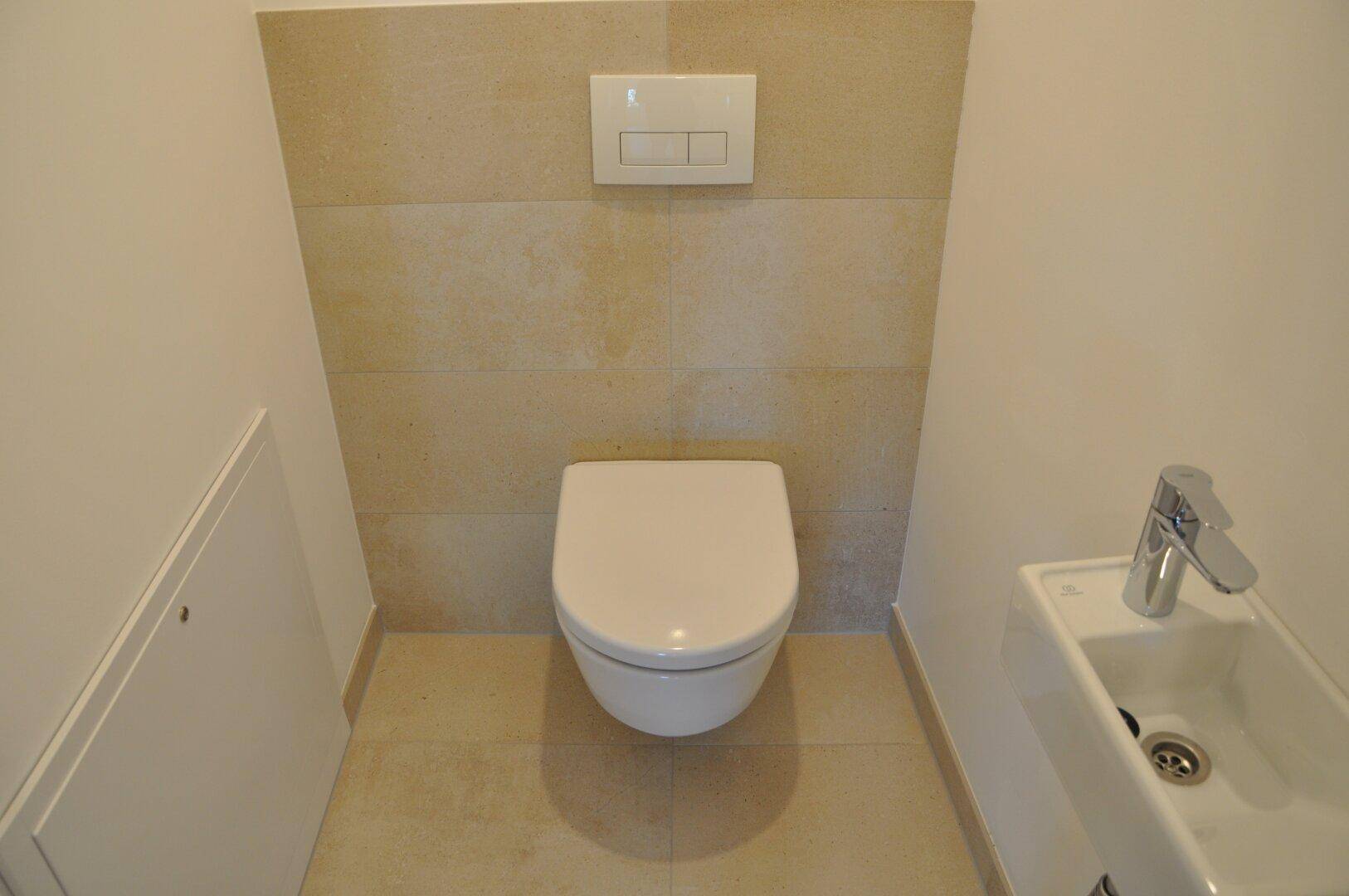Separates WC mit wandhängender Toilette und kleinem Handwaschbecken, modern gefliest.