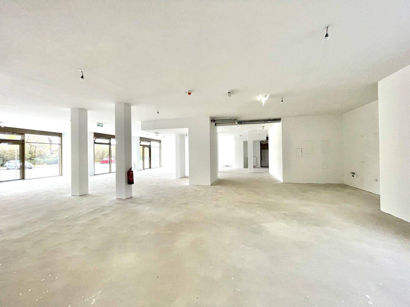 Großer, heller Gewerberaum im Erdgeschoss mit hohen Decken, Betonboden und großen Fenstern zur Straße.