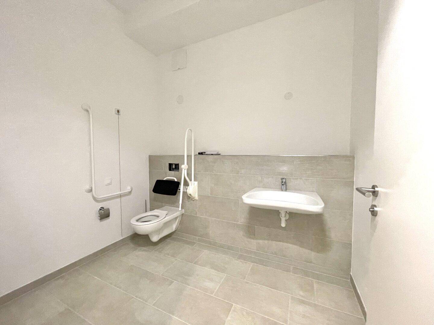 Modernes, barrierefreies Badezimmer mit Wand-WC, Haltegriffen und gefliesten Wänden.