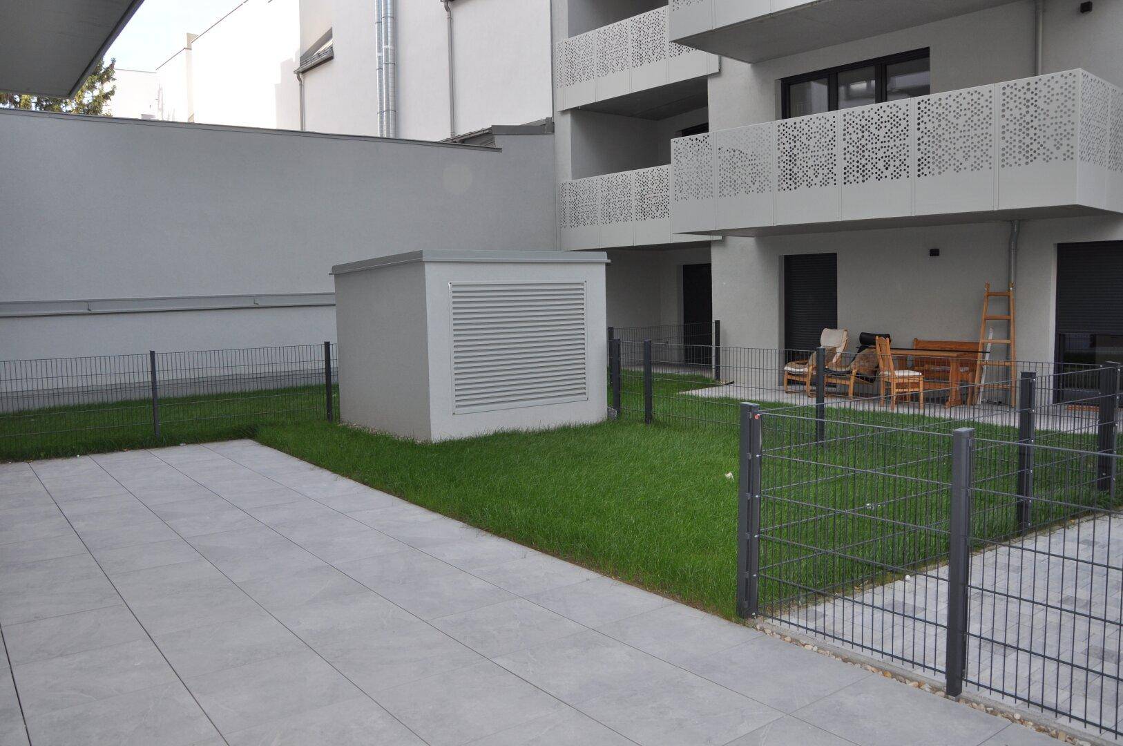 Grünfläche mit Terrasse und einem kleinen Schuppen, angrenzend an moderne Wohnungen.