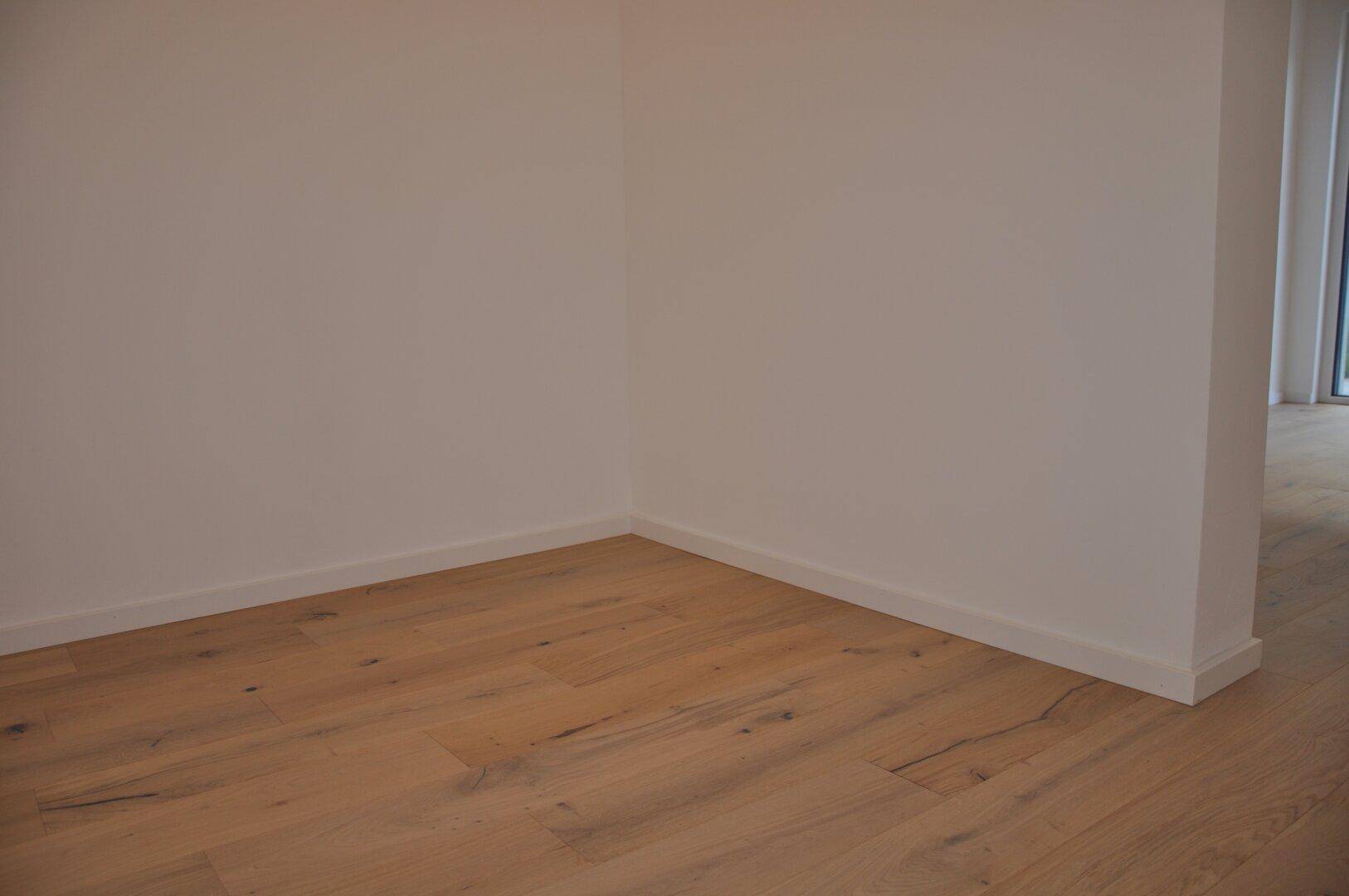 Leere Ecke eines Raumes mit hellem Holzboden und sauberen weißen Wänden.