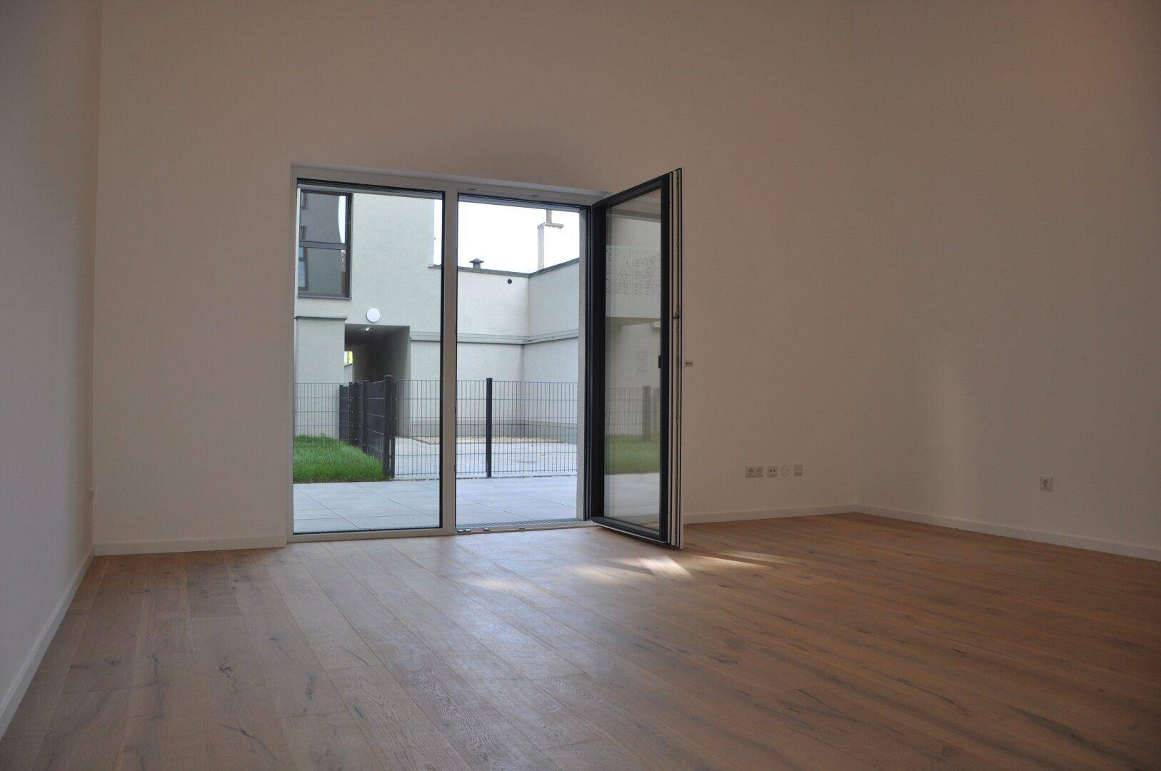 Heller, leerer Raum mit Holzboden und großen Glastüren, die zum Außenbereich führen.