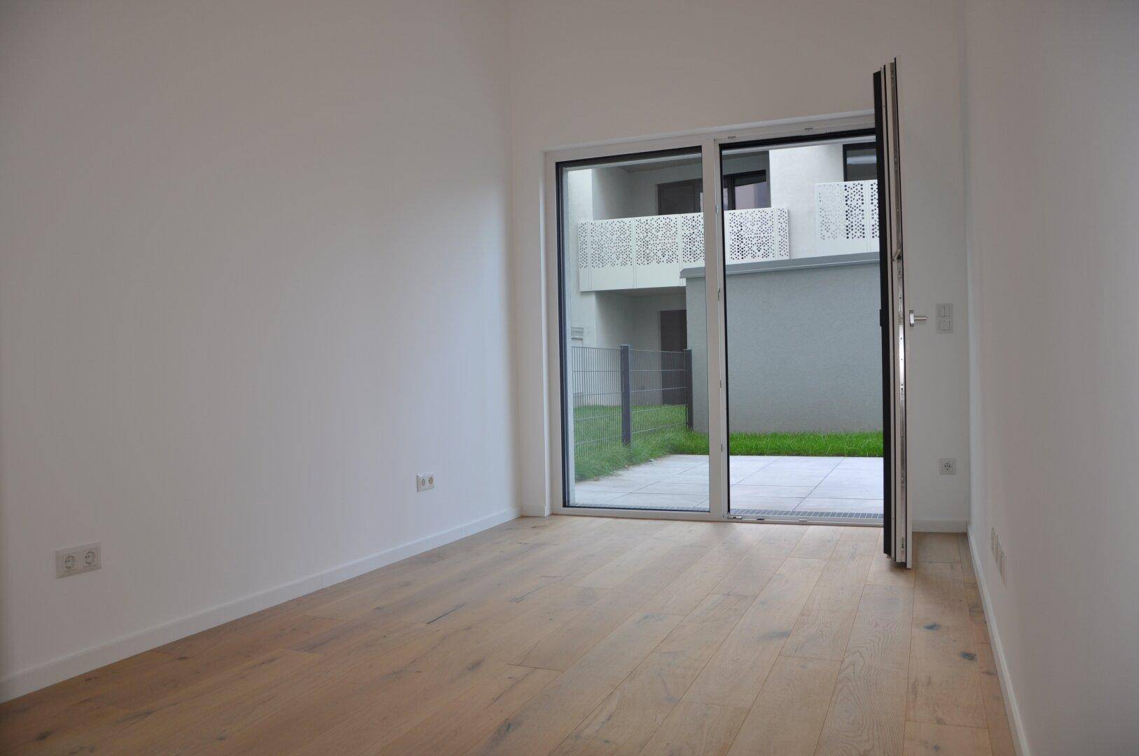 Heller Raum mit Holzboden und Glastür, die Zugang zu einem Außenbereich bietet.