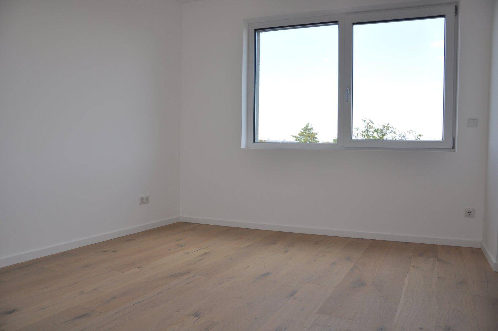 Helles Schlafzimmer mit Holzboden und Fenster, das einen Blick ins Grüne bietet.