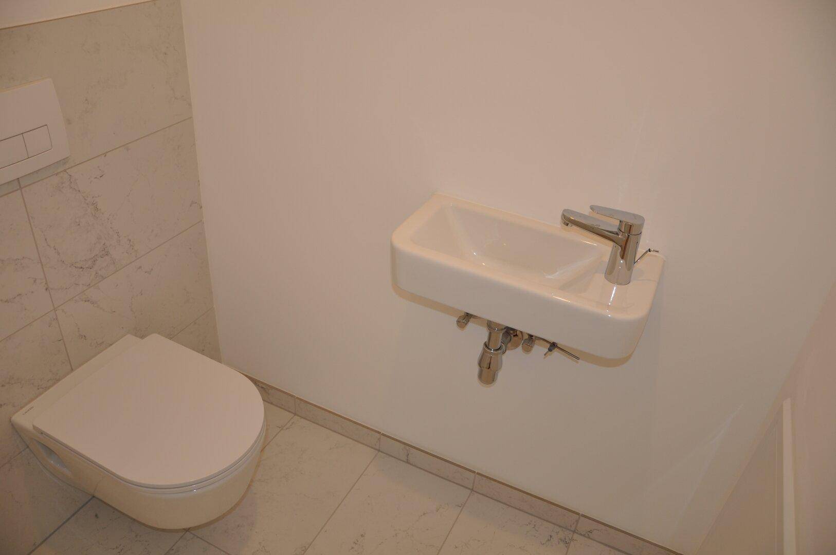 Separates WC mit wandhängender Toilette und kleinem Waschbecken, modern gefliest.
