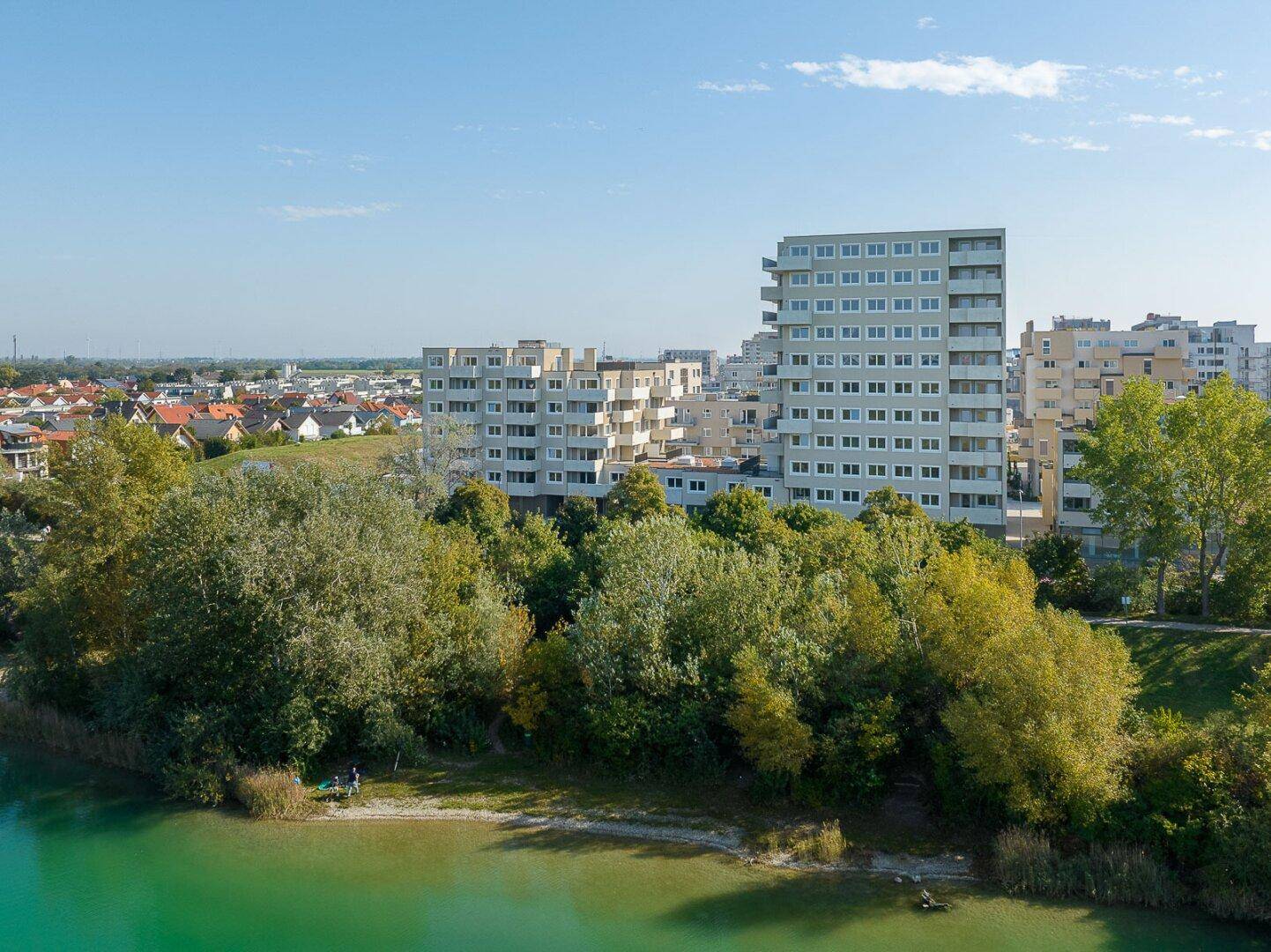 Weitläufiger Blick auf einen türkisfarbenen See mit bewaldetem Ufer und Wohngebäuden im Hintergrund unter blauem Himmel.