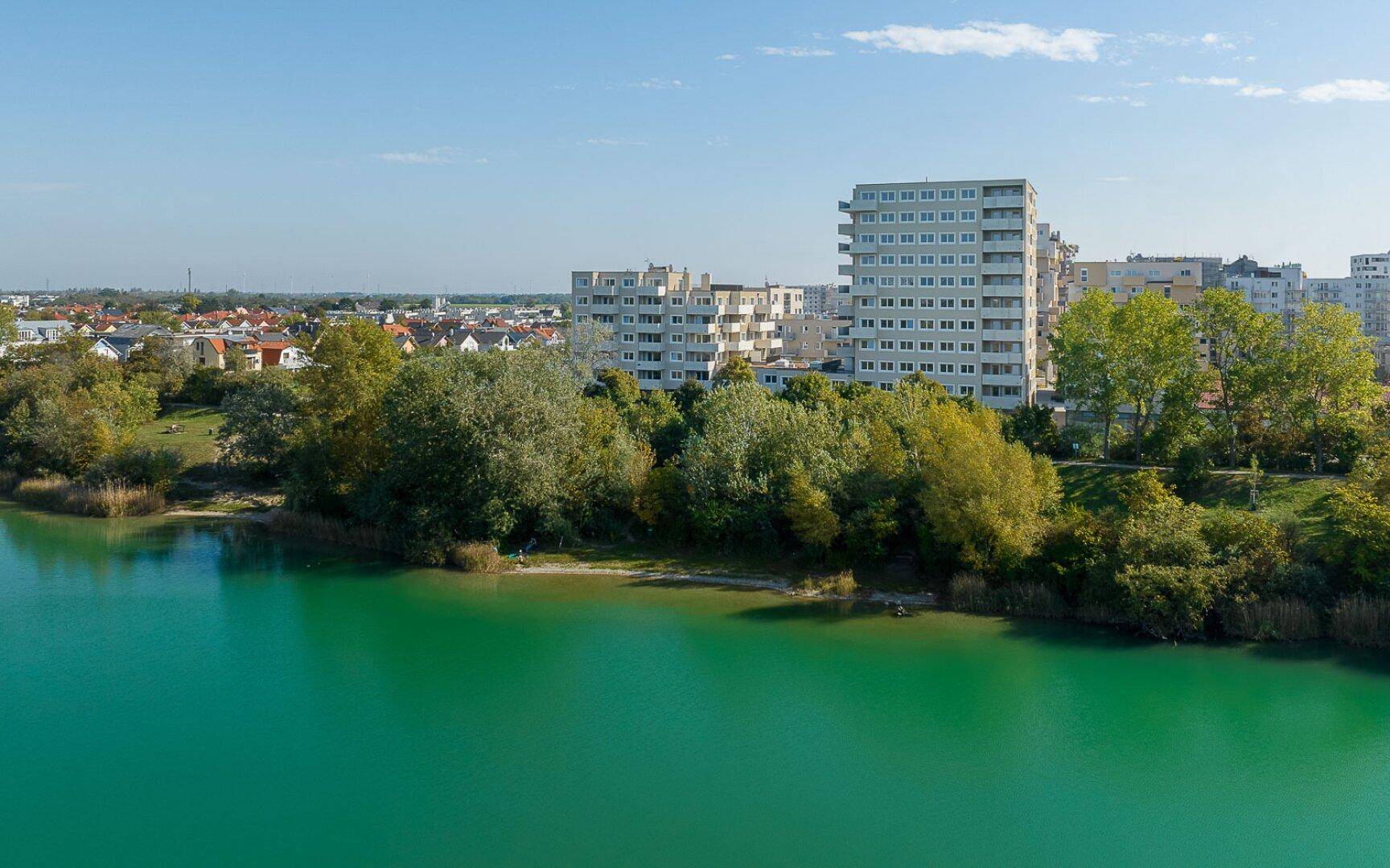 Panoramablick auf einen ruhigen See mit üppiger Vegetation am Ufer und modernen Wohnkomplexen in der Ferne.