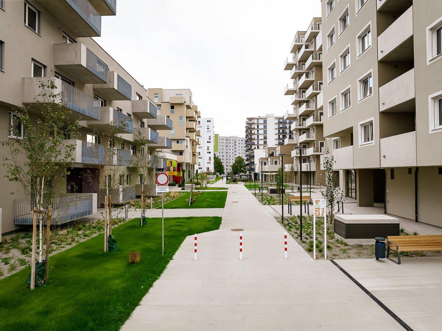 Fußgängerweg zwischen modernen Wohngebäuden mit gepflegten Grünstreifen und Sitzbänken.