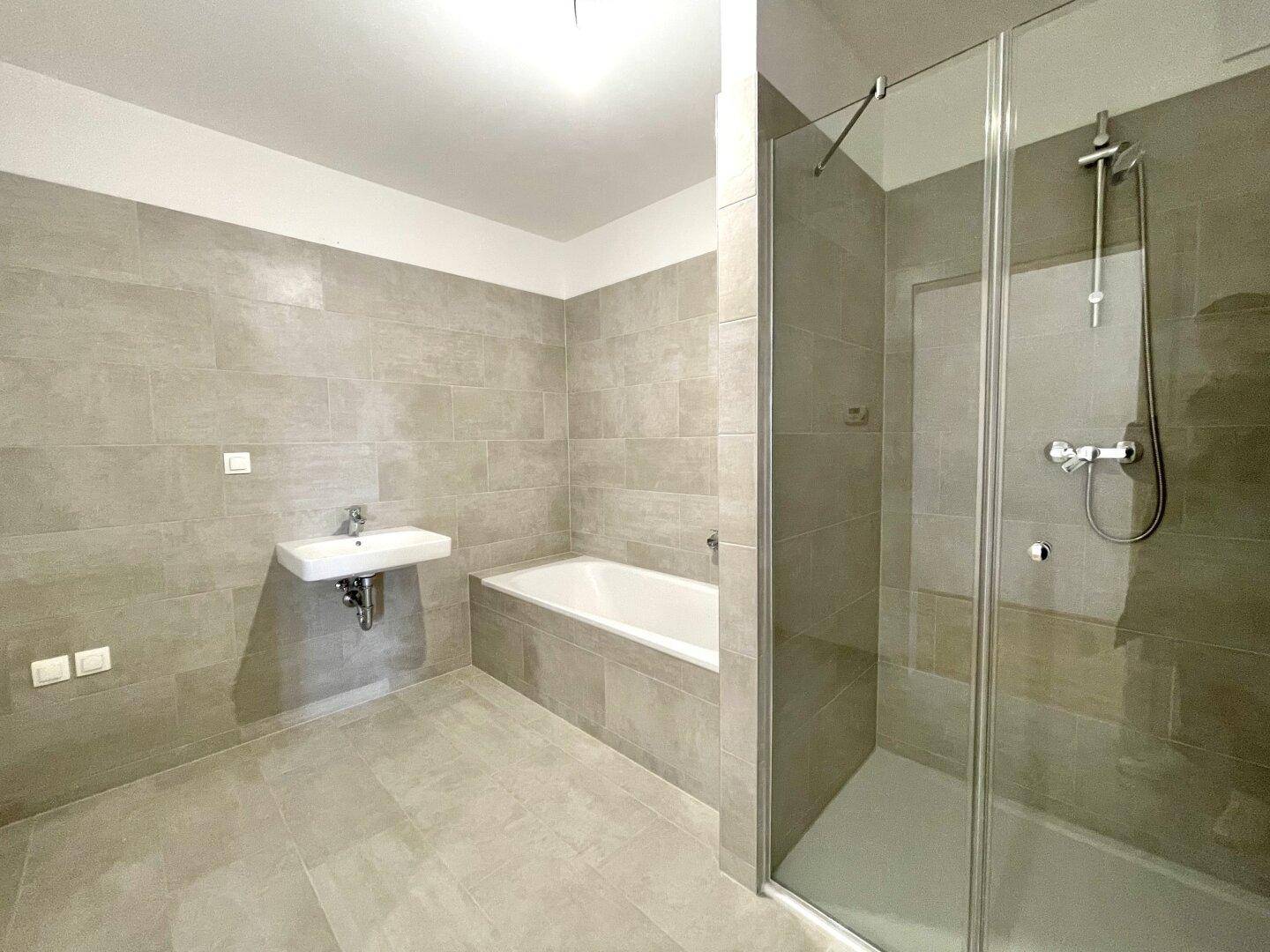 Modernes Badezimmer mit grauen Fliesen, Badewanne, Waschbecken und einer Duschkabine aus Glas.