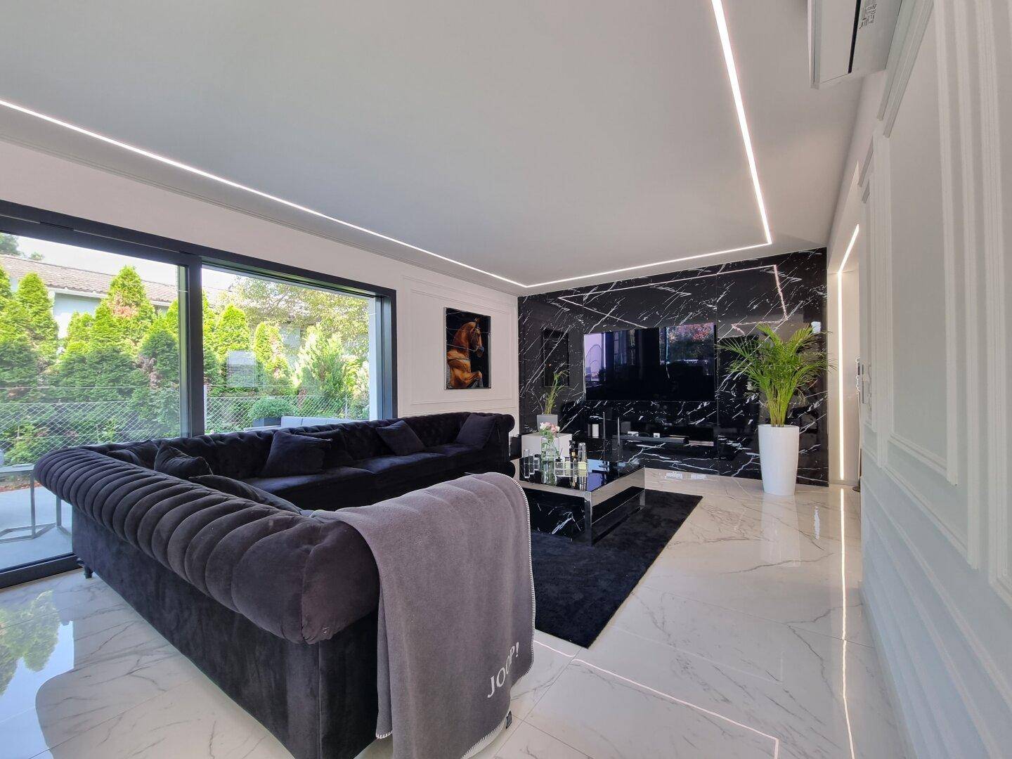 Elegantes Wohnzimmer mit schwarzem Sofa, Marmorwand, großen Fenstern und modernen Deckenleuchten.
