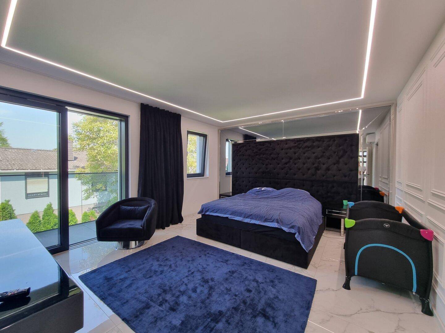 Geräumiges Schlafzimmer mit großem schwarzem Bett, blauem Teppich und Zugang zum Balkon.