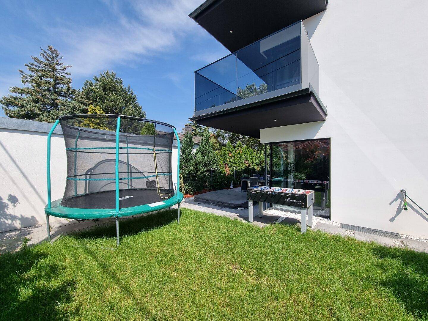 Gartenbereich mit Trampolin und Tischfußball vor der modernen Hausfassade mit Glasbalkon.